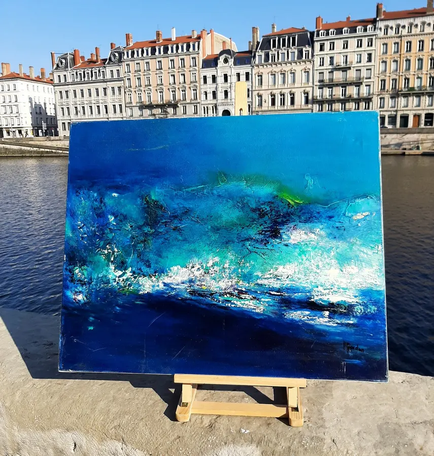 Tableau abstrait bleu de Régine Gardan, touches turquoise et blanches, sur un chevalet au bord d'un fleuve, devant des façades anciennes
